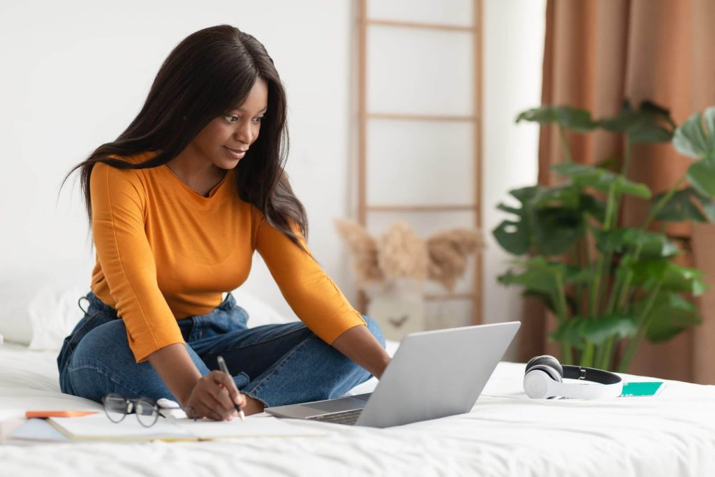 Black Woman Taking Notes Sitting At Laptop Learning At Home | Govcon